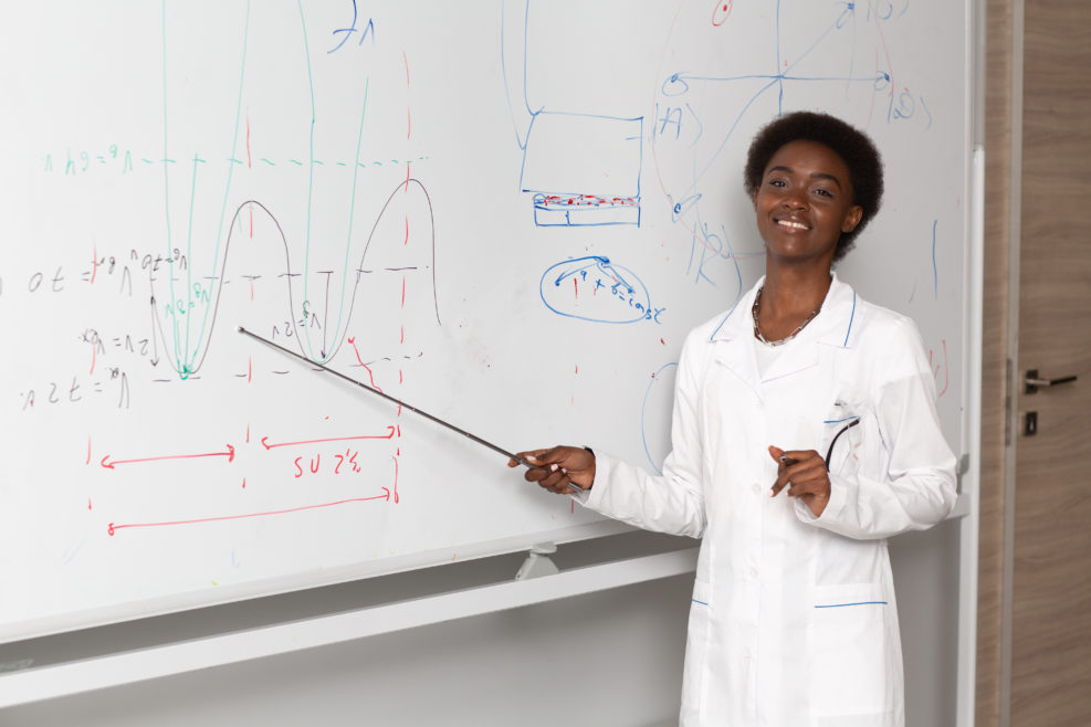 African American smiling woman math teacher stands at black board with pointer.